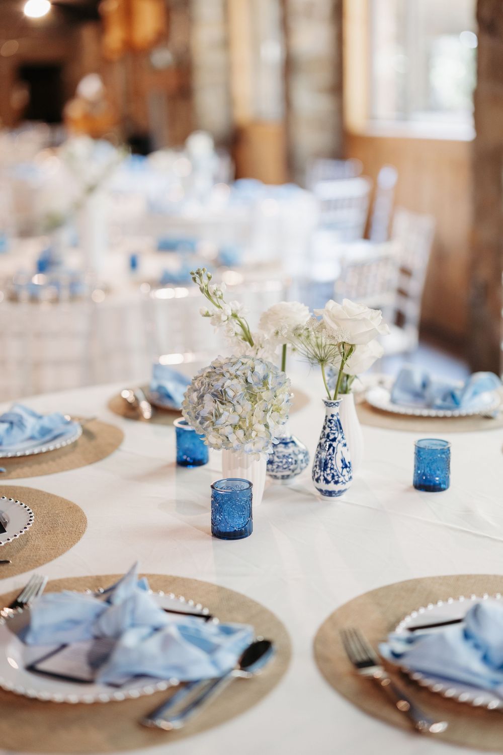 Elegant wedding table setting with blue napkins, white floral centerpieces, blue glass votives, and woven charger plates.