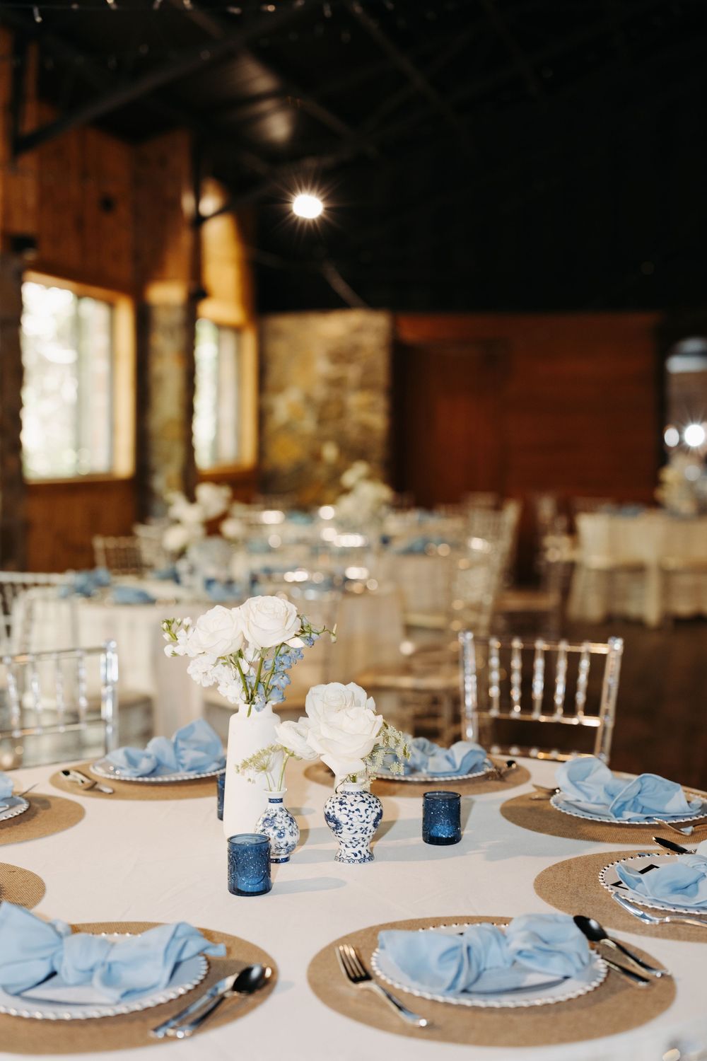 Elegant wedding reception table with blue napkins, white roses, and blue candle holders in a rustic barn venue.