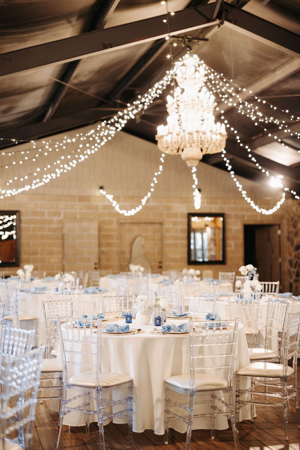 Elegant wedding reception hall with fairy lights, chandelier, white round tables, blue accents, and clear chiavari chairs.