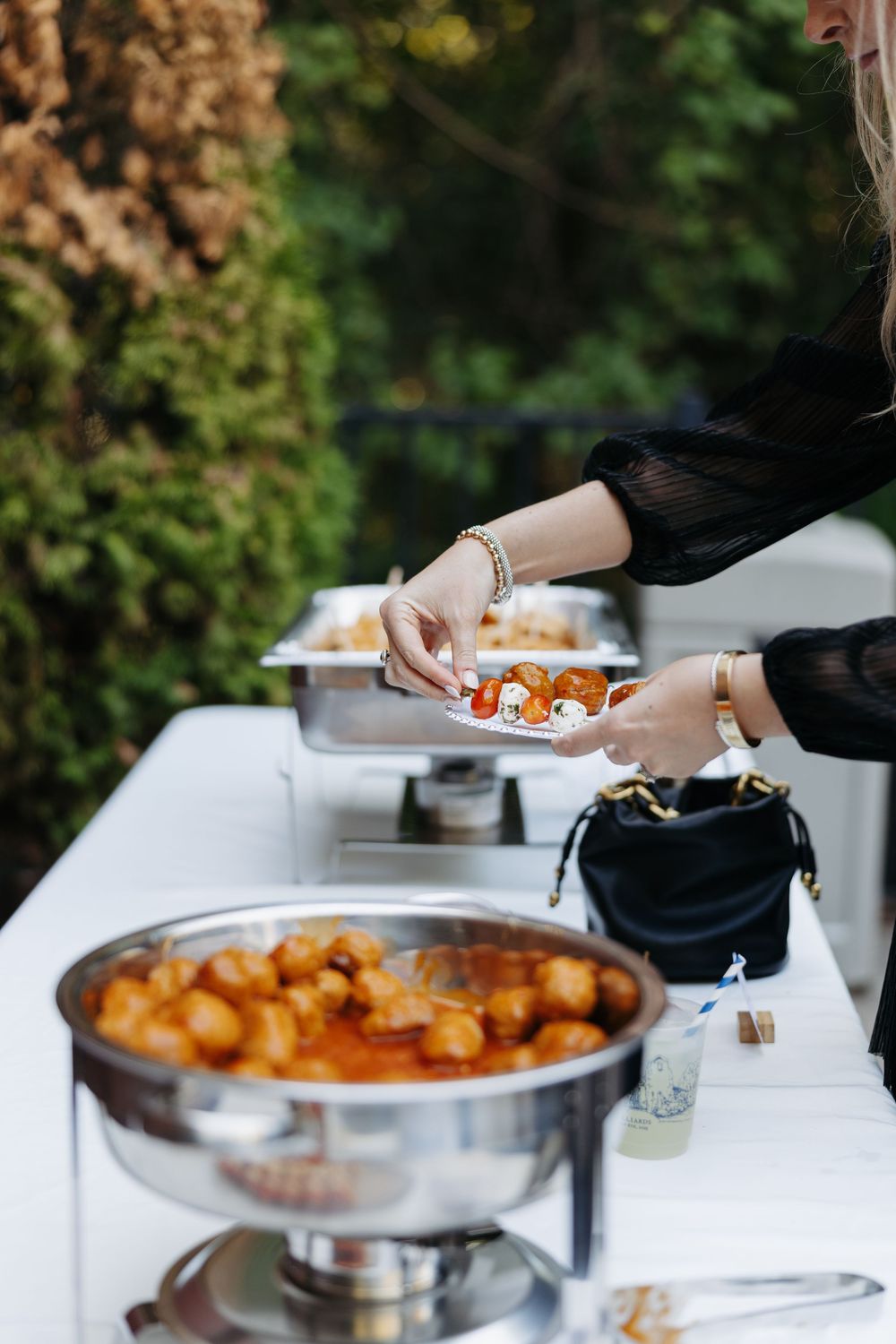Woman serving food at an elegant outdoor buffet with chafing dishes filled with saucy appetizers at a catered event.