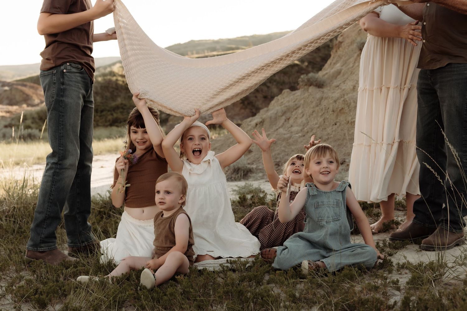 Five young children sit outdoors under a flowing fabric held by adults in a scenic natural setting.