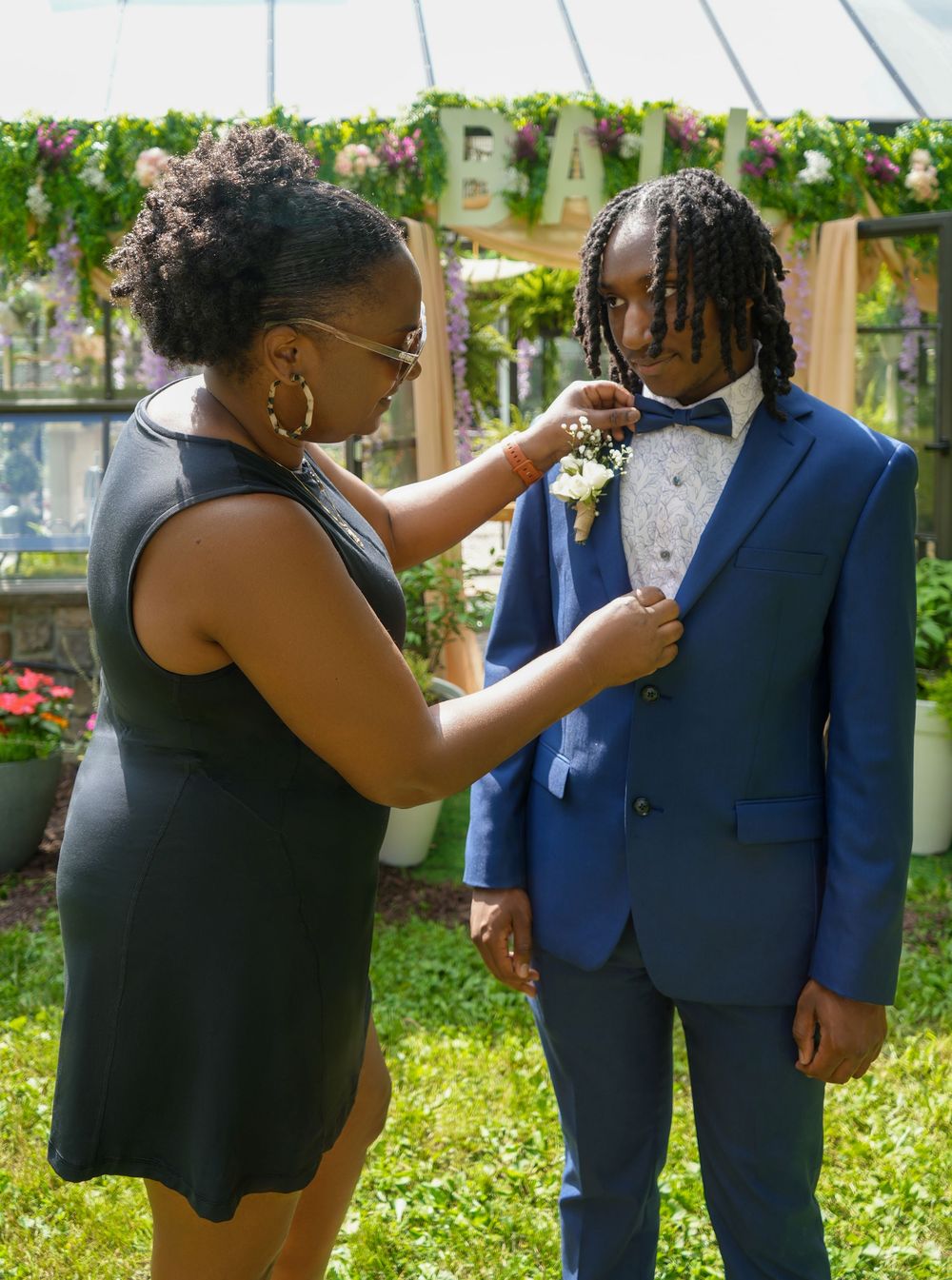 A woman pins a boutonniere on a young man in a blue suit and bow tie outdoors at a formal event.