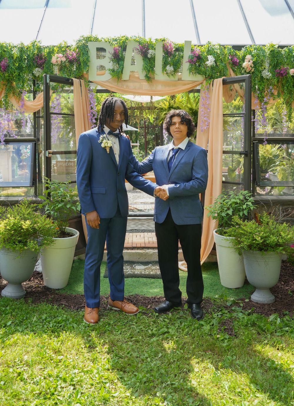 Two young men in blue suits shake hands under a floral BALL arch outdoors.