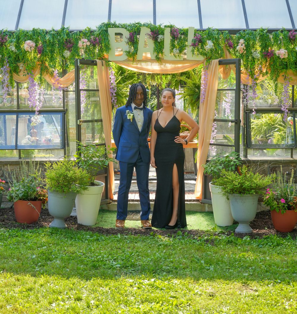 Couple in formal attire posing under floral arch decorated with BALL sign at prom venue.