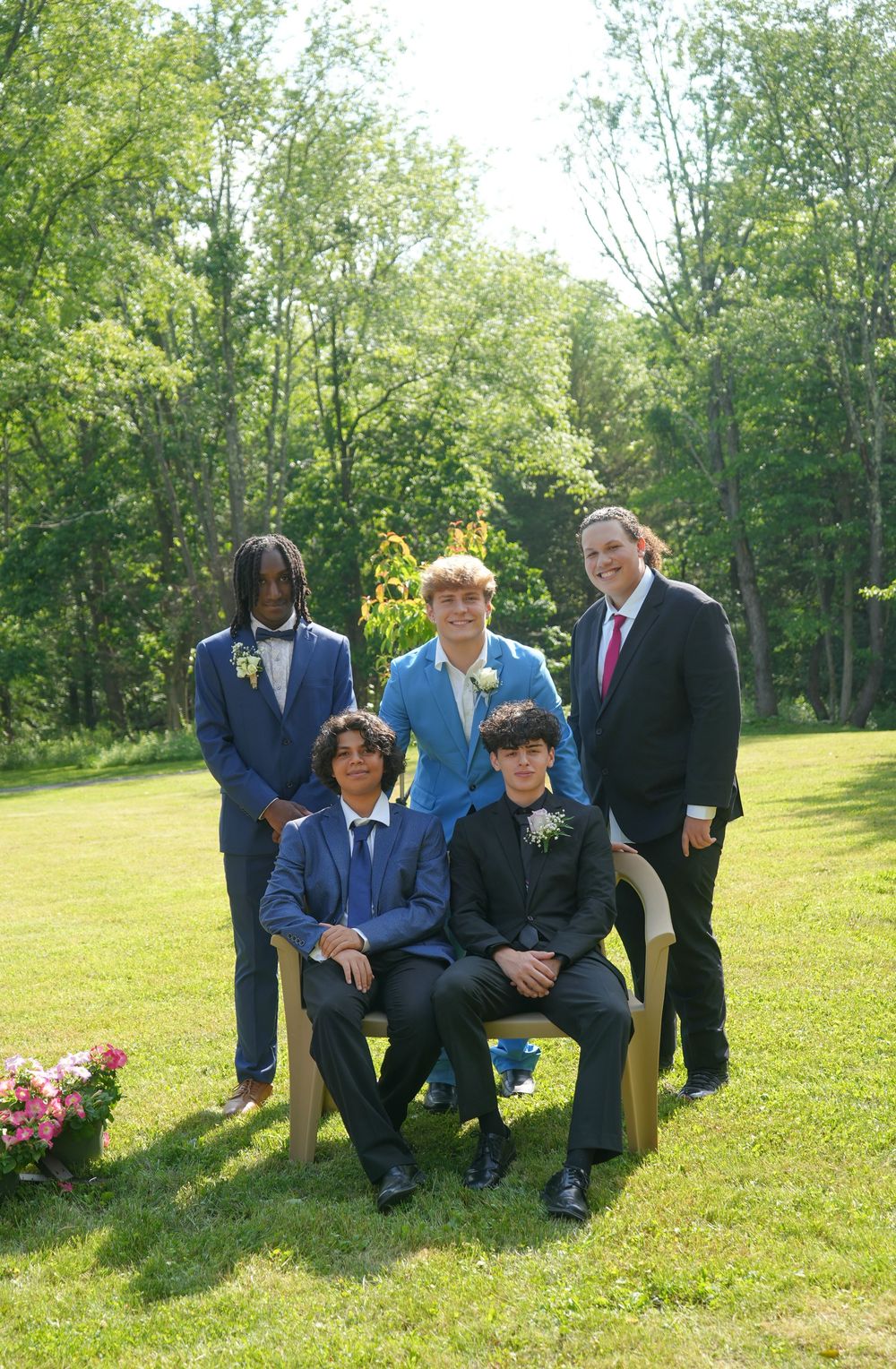 Five young men in formal suits pose outdoors on a sunny day, two seated in chairs on a green lawn.