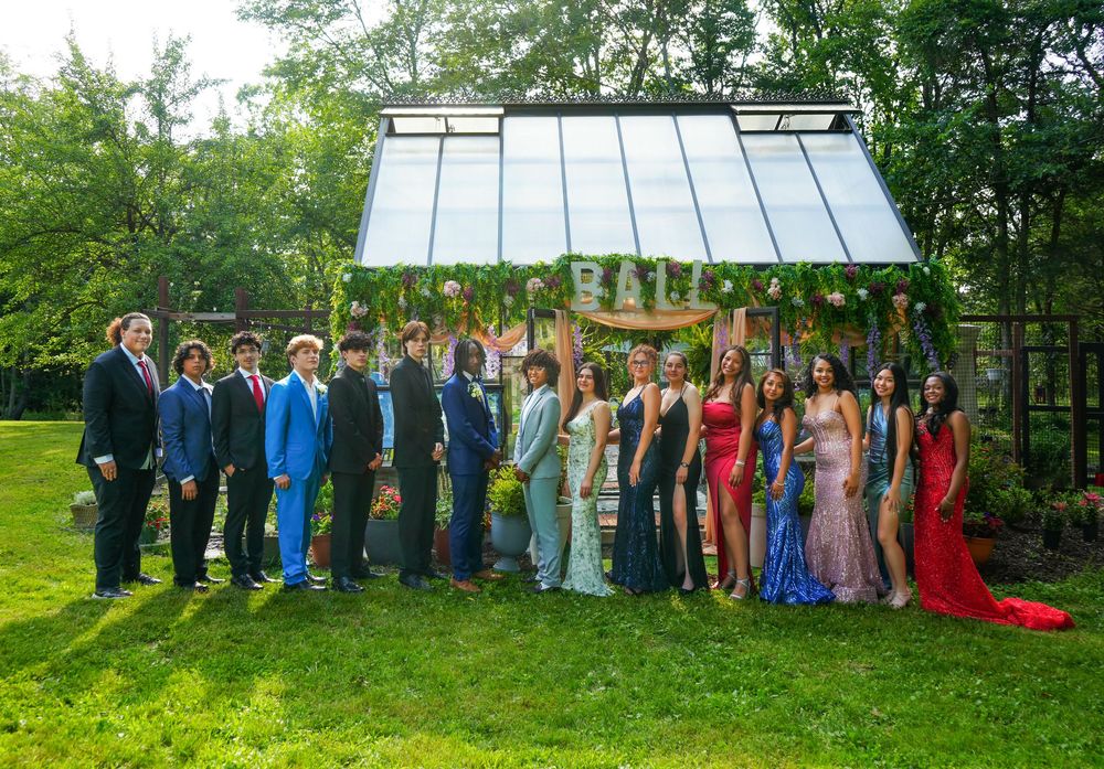 Group of teenagers in formal prom attire posing outdoors in front of a decorated greenhouse backdrop.