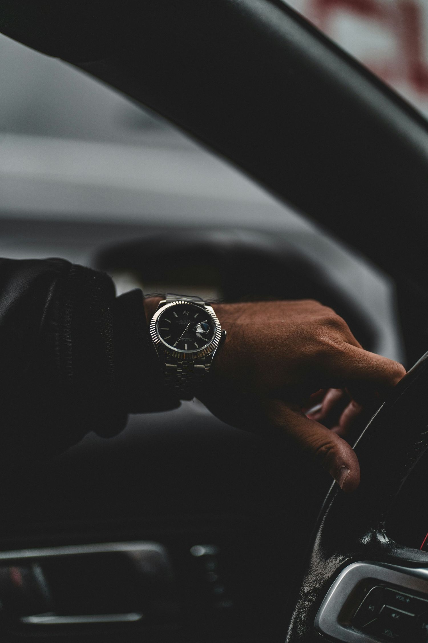 A hand wearing a silver luxury watch grips a car steering wheel, showcasing elegant style while driving.