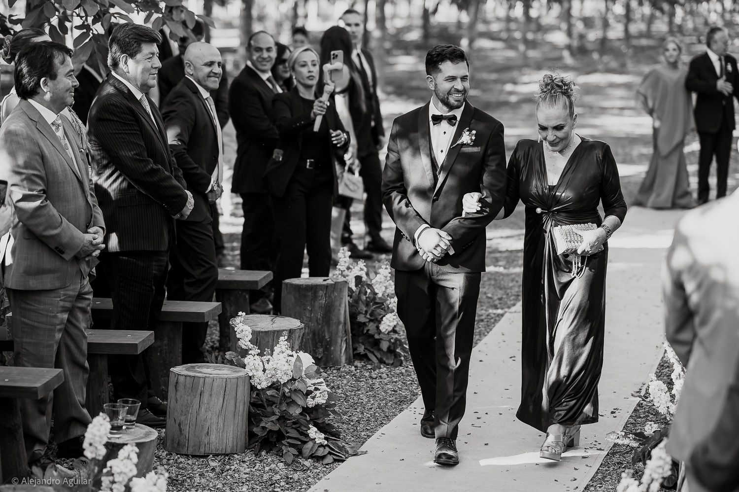 Groom walks down outdoor wedding aisle with elegantly dressed woman, guests watching from rustic log benches.