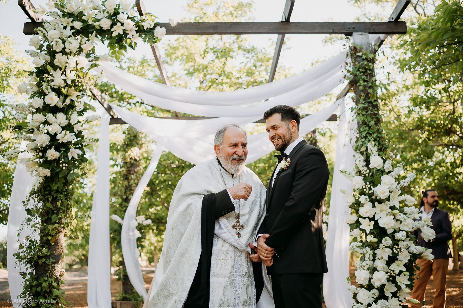 Groom smiling with officiant under floral wedding arch adorned with white flowers and draped fabric outdoors.