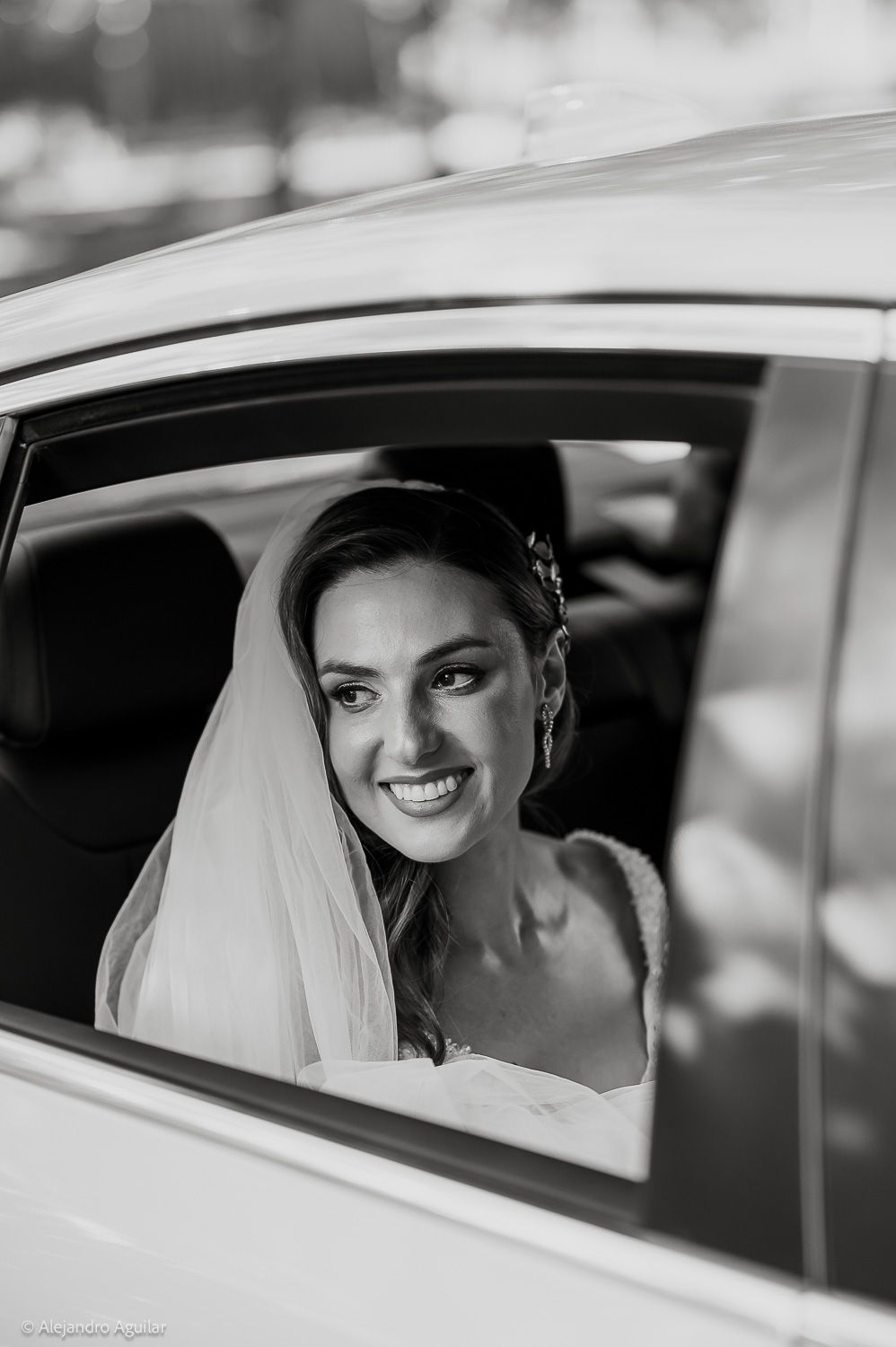 Black and white portrait of a smiling bride in a veil looking out of a car window on her wedding day.