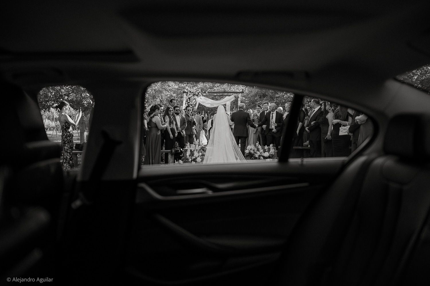 Black and white wedding photo shot from inside a car, capturing bride walking down the aisle through the window.