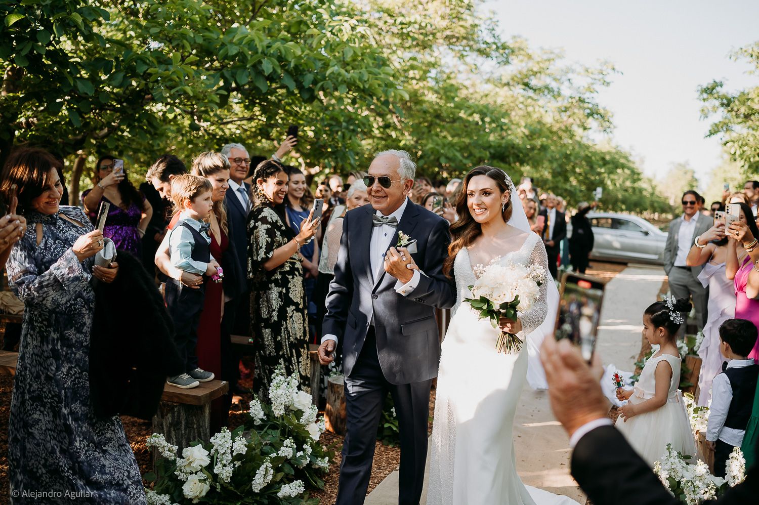 Bride walks down outdoor aisle with escort, guests applauding, surrounded by trees and white floral arrangements.