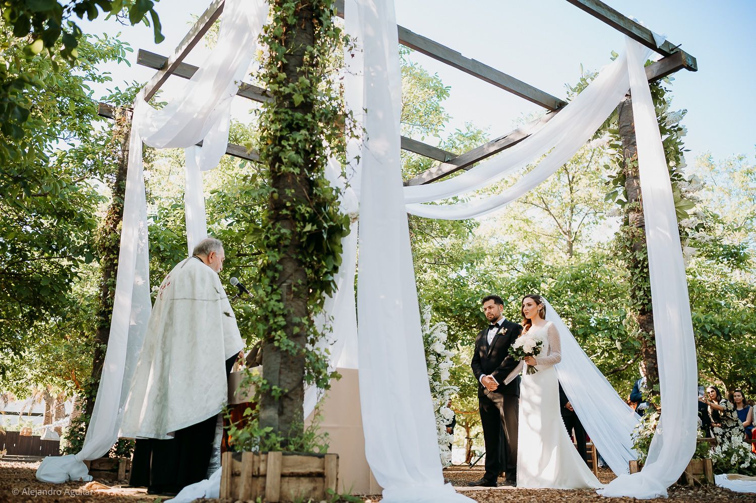Outdoor wedding ceremony under a rustic wooden arbor draped with white fabric and greenery, bride and groom with officiant.