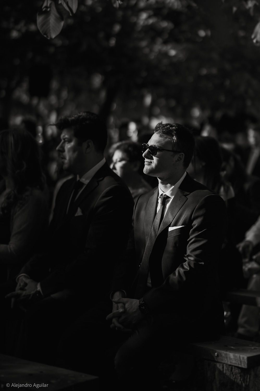 Black and white photo of a smiling man in a suit and sunglasses seated at an outdoor event.