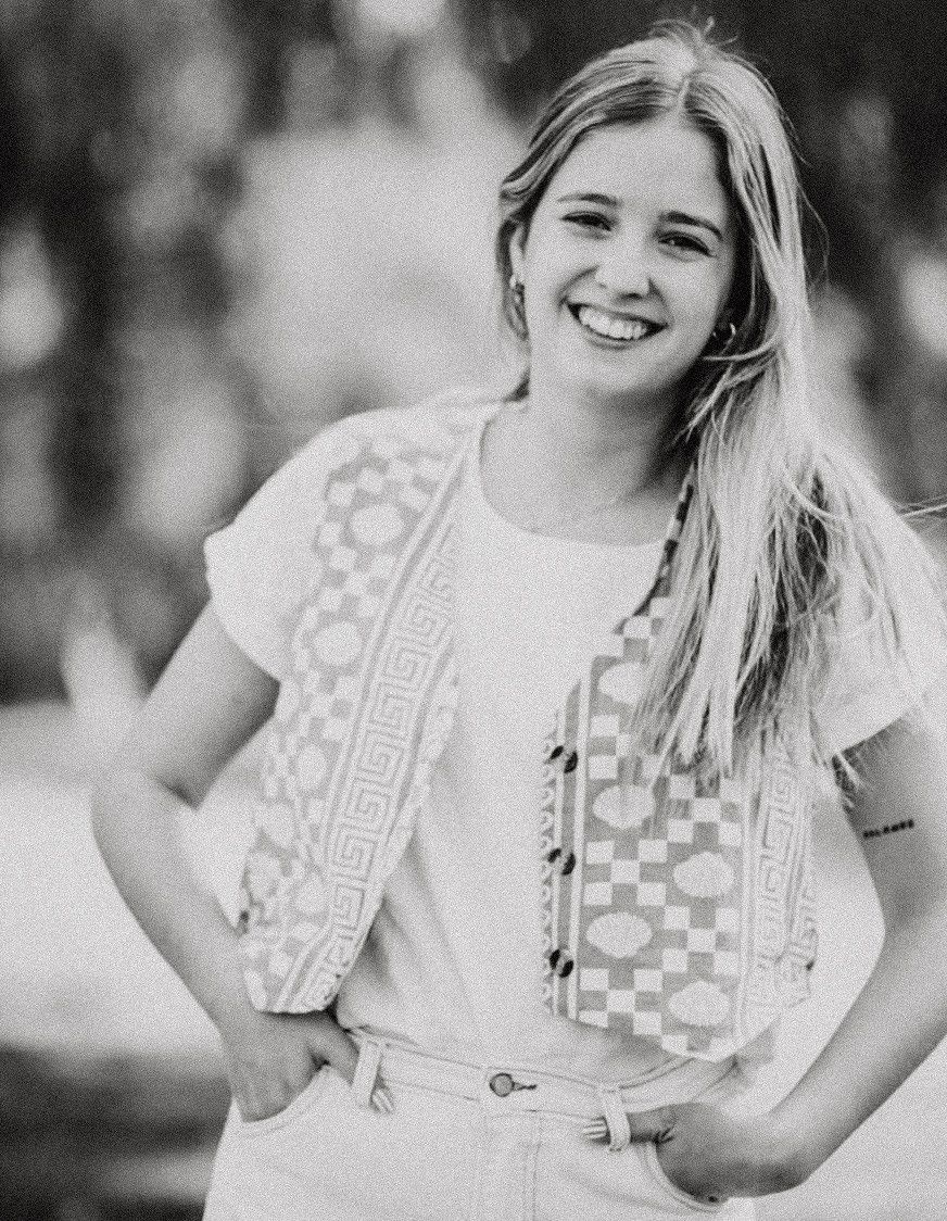 Smiling young woman with long blonde hair wearing a patterned vest and white top, posing outdoors in a black and white photo.