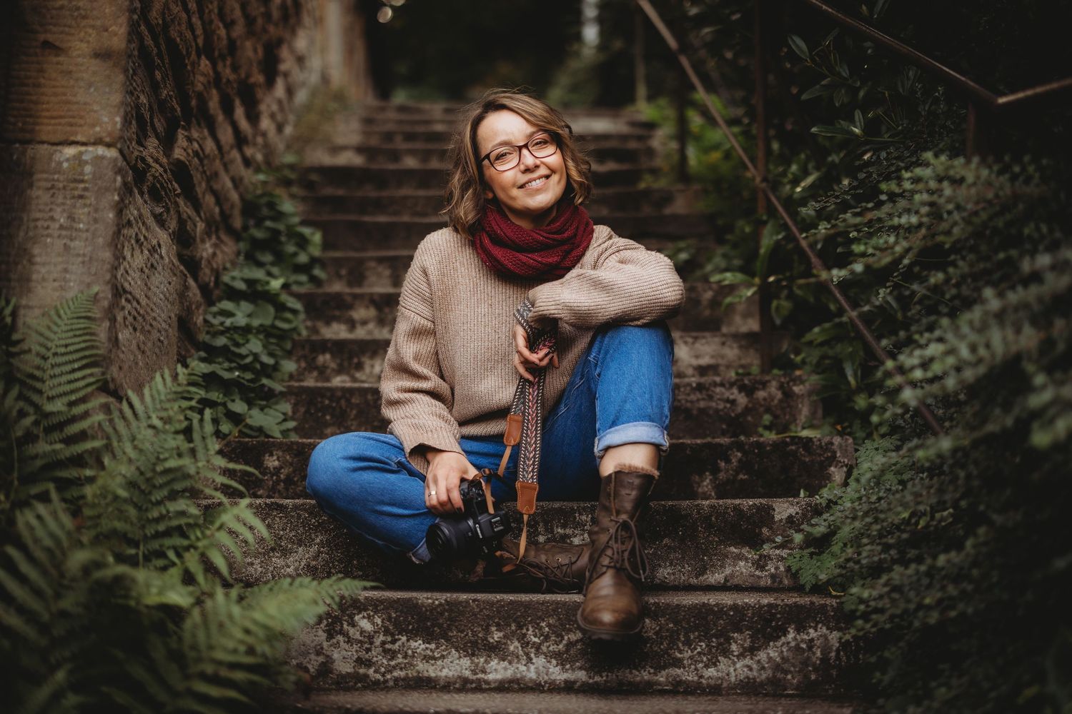 Smiling woman with short blonde hair in a knit sweater posing outdoors in a sunlit forest with a dog behind her.