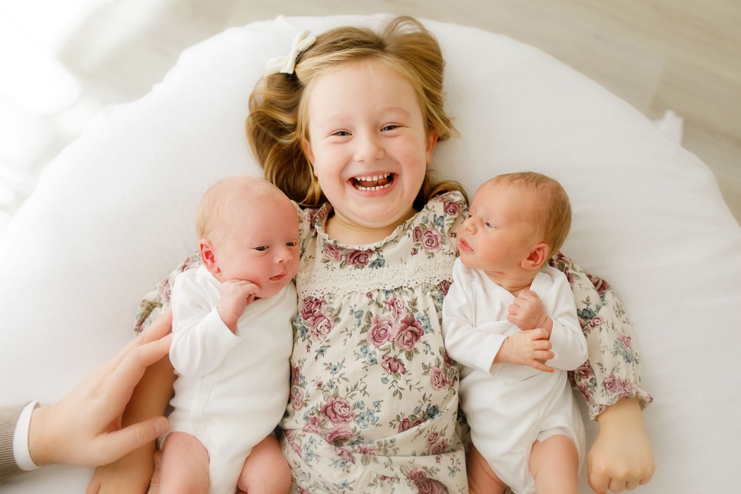 Laughing young girl in floral dress lying with twin newborn babies in white onesies on a white bed.