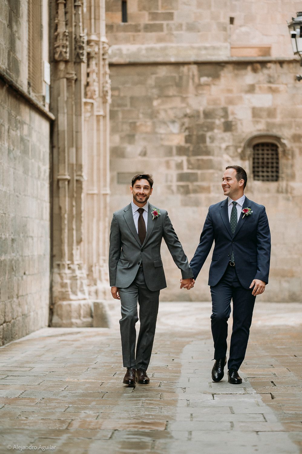 Two grooms in suits hold hands while walking past a historic stone cathedral on their wedding day.