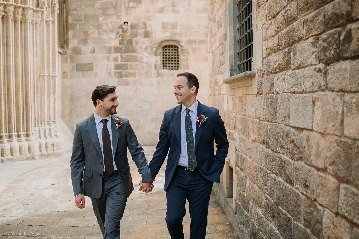 Two grooms hold hands and smile while walking past a stone building on their wedding day.