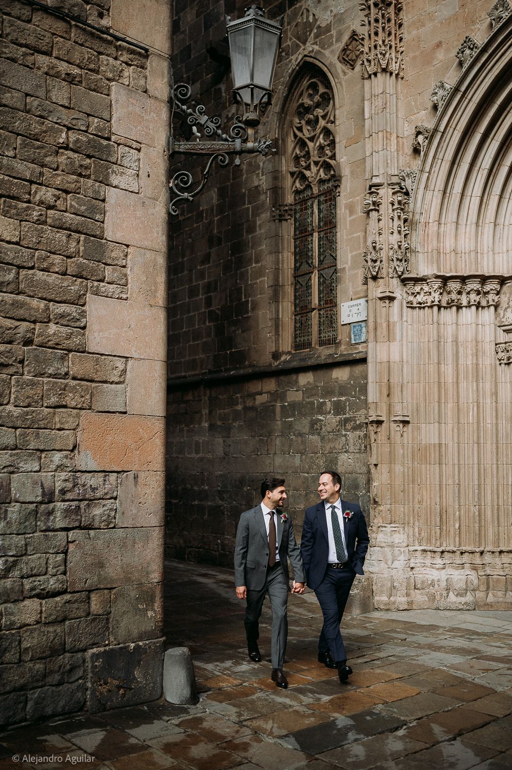Two grooms in suits hold hands walking past a Gothic cathedral in Barcelona's historic quarter.