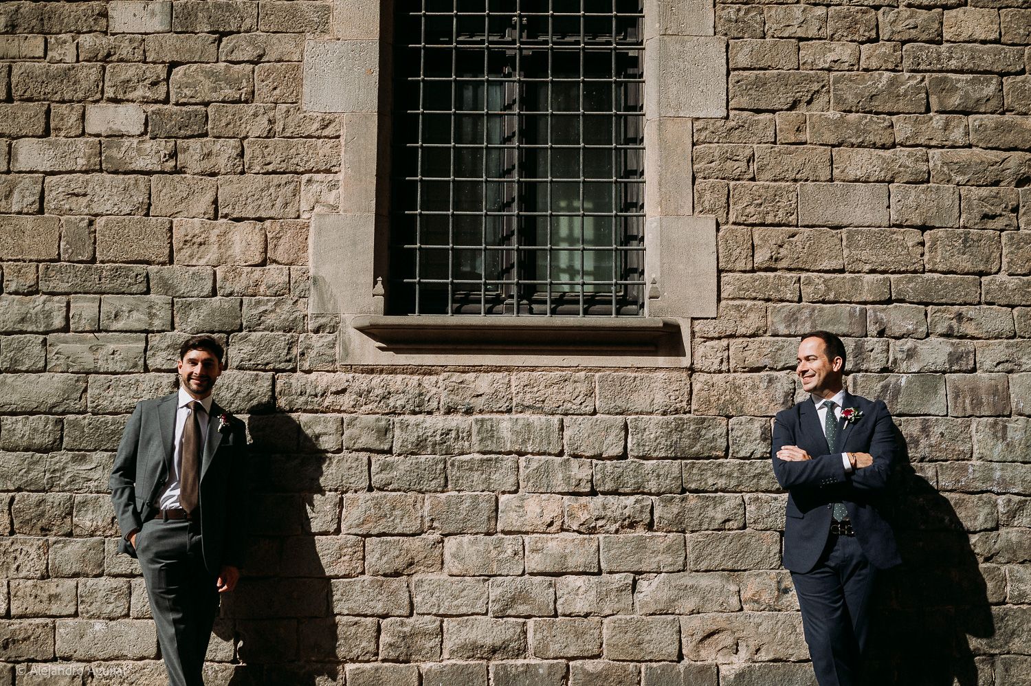 Two men in suits stand against a rustic stone wall beneath a barred window in natural sunlight.