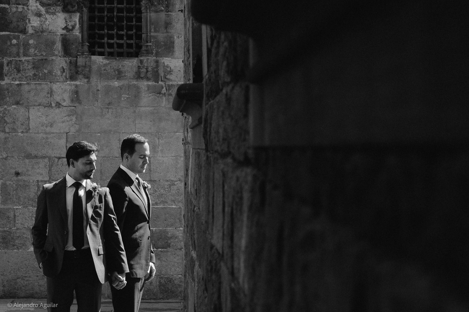 Two grooms in suits share an intimate moment against a stone wall in a black and white wedding photo.