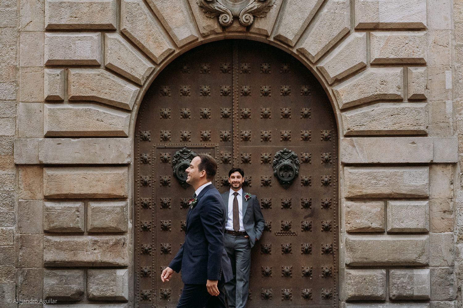 Two grooms in suits stand before an ornate wooden door with iron studs on a stone building facade.
