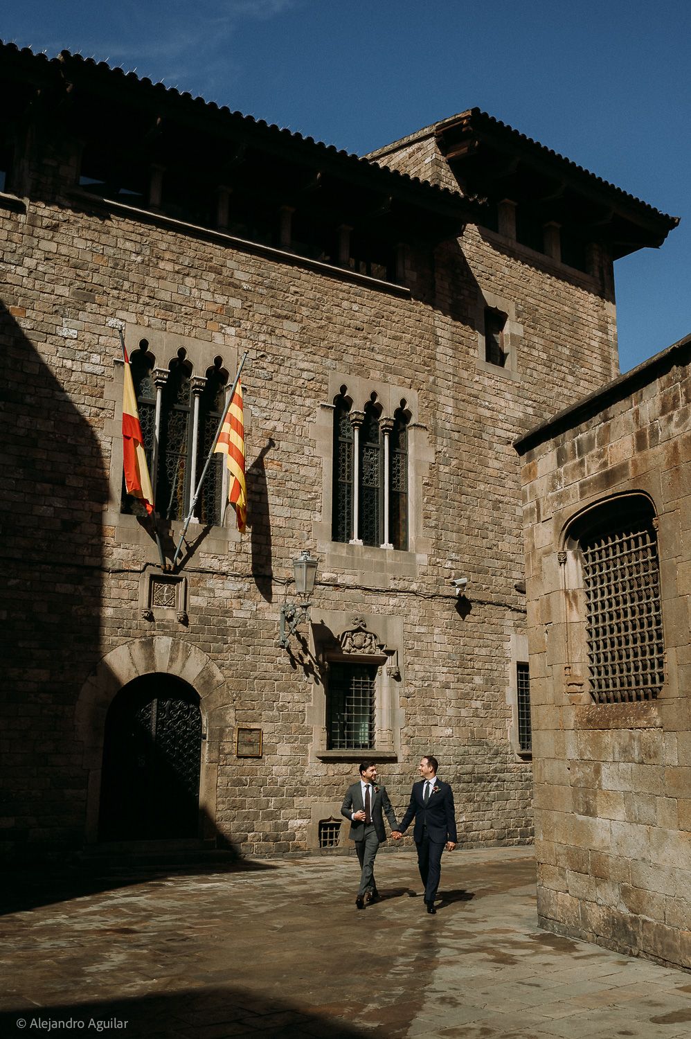 Two grooms in suits hold hands walking past a historic stone building with flags in Barcelona, Spain.