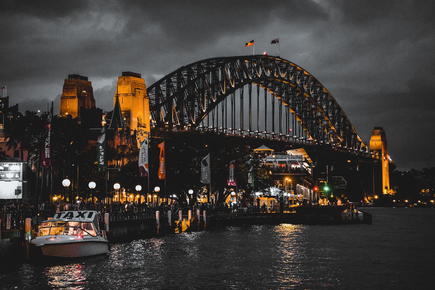 Sydney Harbour Bridge illuminated at night with boats on dark water under cloudy sky.