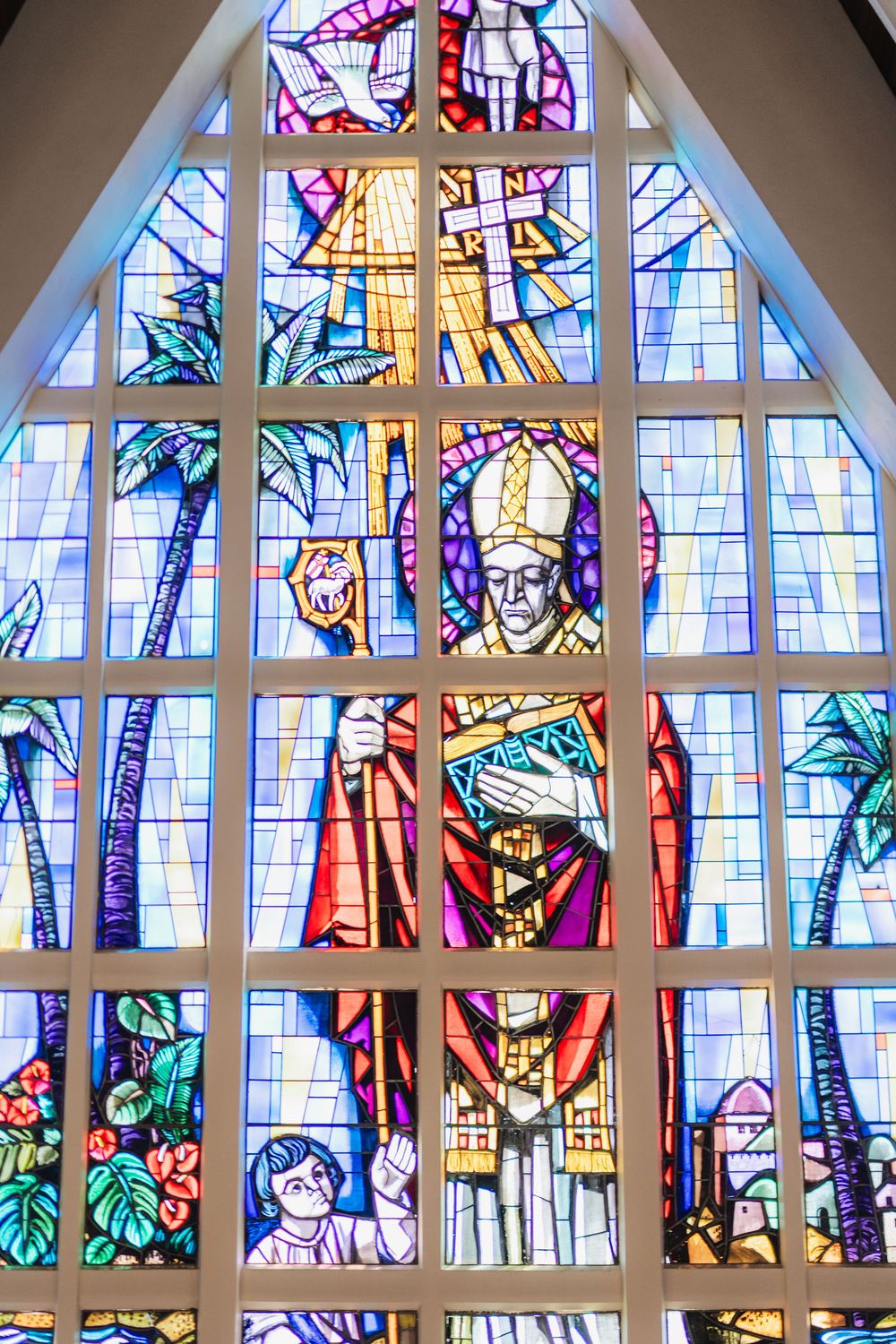 Colorful stained glass window in a church depicting a bishop in robes holding a cross, with palm trees and floral motifs.