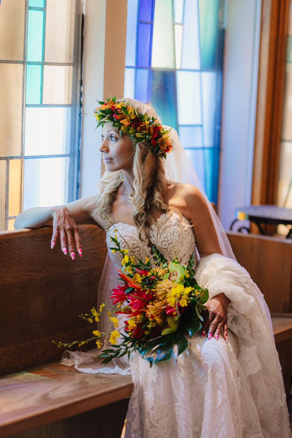 Elegant bride in lace gown with colorful floral crown holds vibrant bouquet beside stained glass church window.