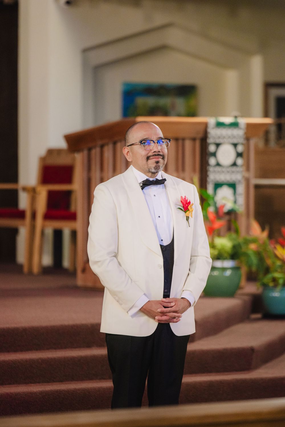 Groom in white tuxedo jacket with floral boutonniere stands at church altar awaiting bride during wedding ceremony.