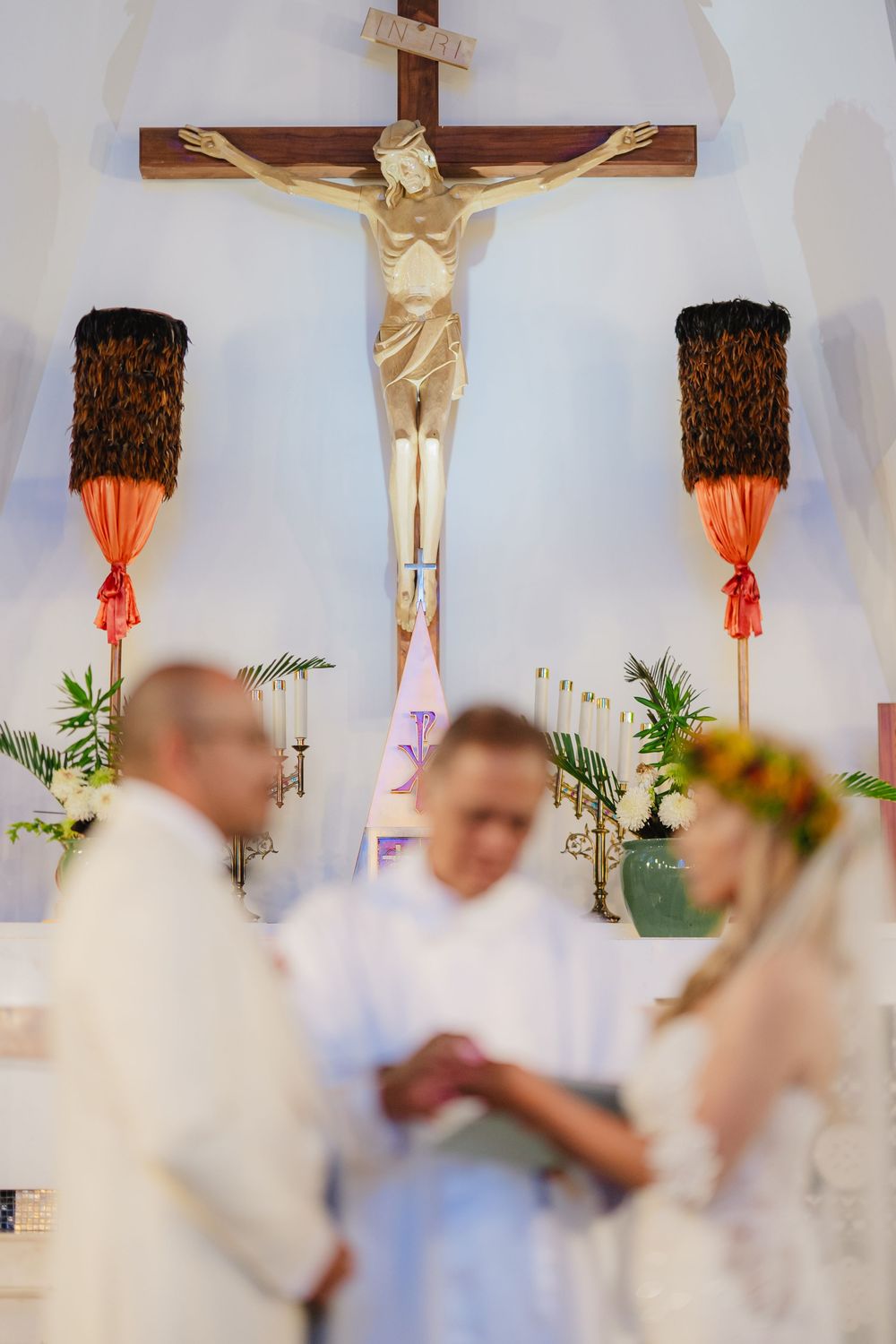 Catholic wedding ceremony with couple exchanging vows before a crucifix, officiated by a priest in a white chapel.