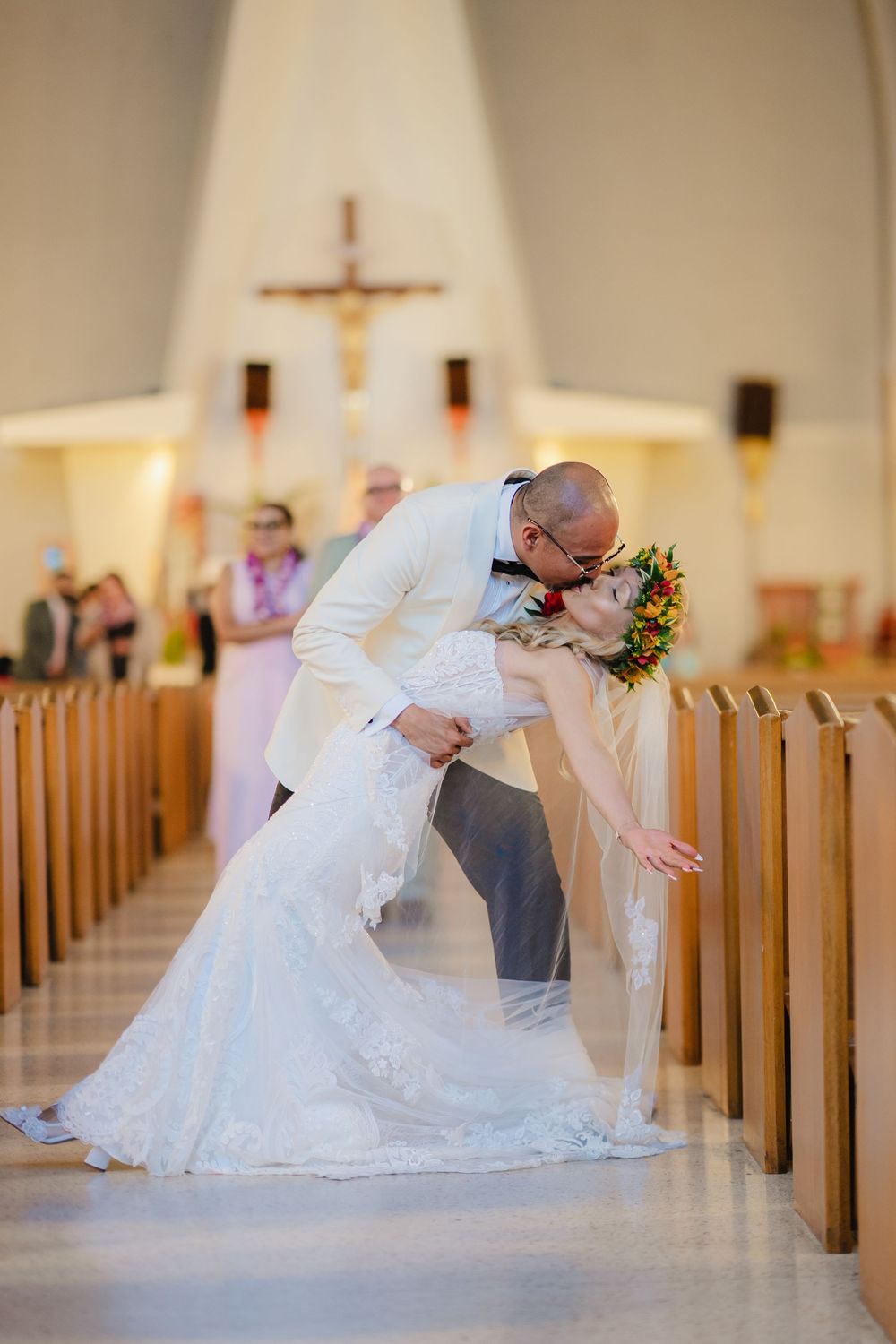 Groom dips bride for a kiss in church aisle after wedding ceremony, with cross altar and guests in background.