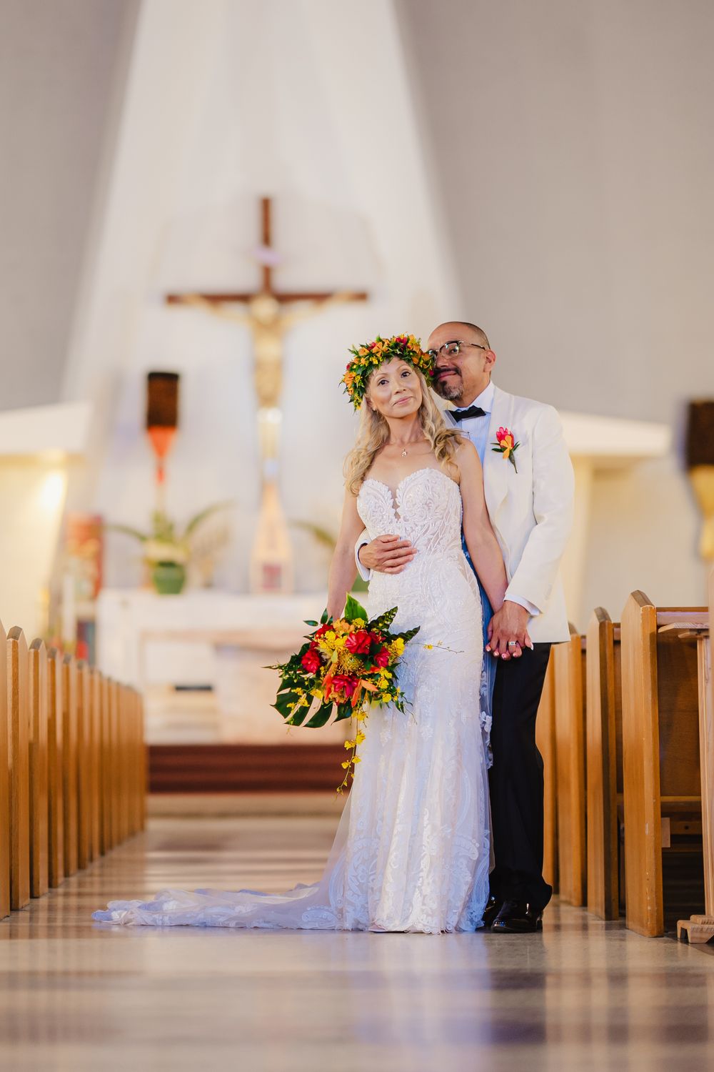 Bride in white lace gown and floral crown stands with groom in white jacket inside a church aisle on their wedding day.