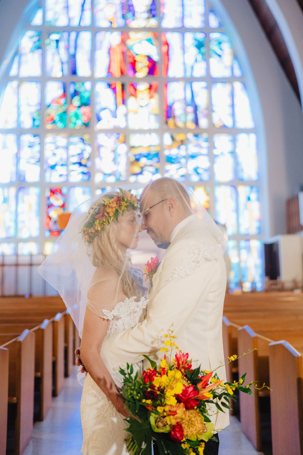 Bride and groom share a romantic moment in a church with a stunning stained glass window backdrop, bride holding colorful bouquet.