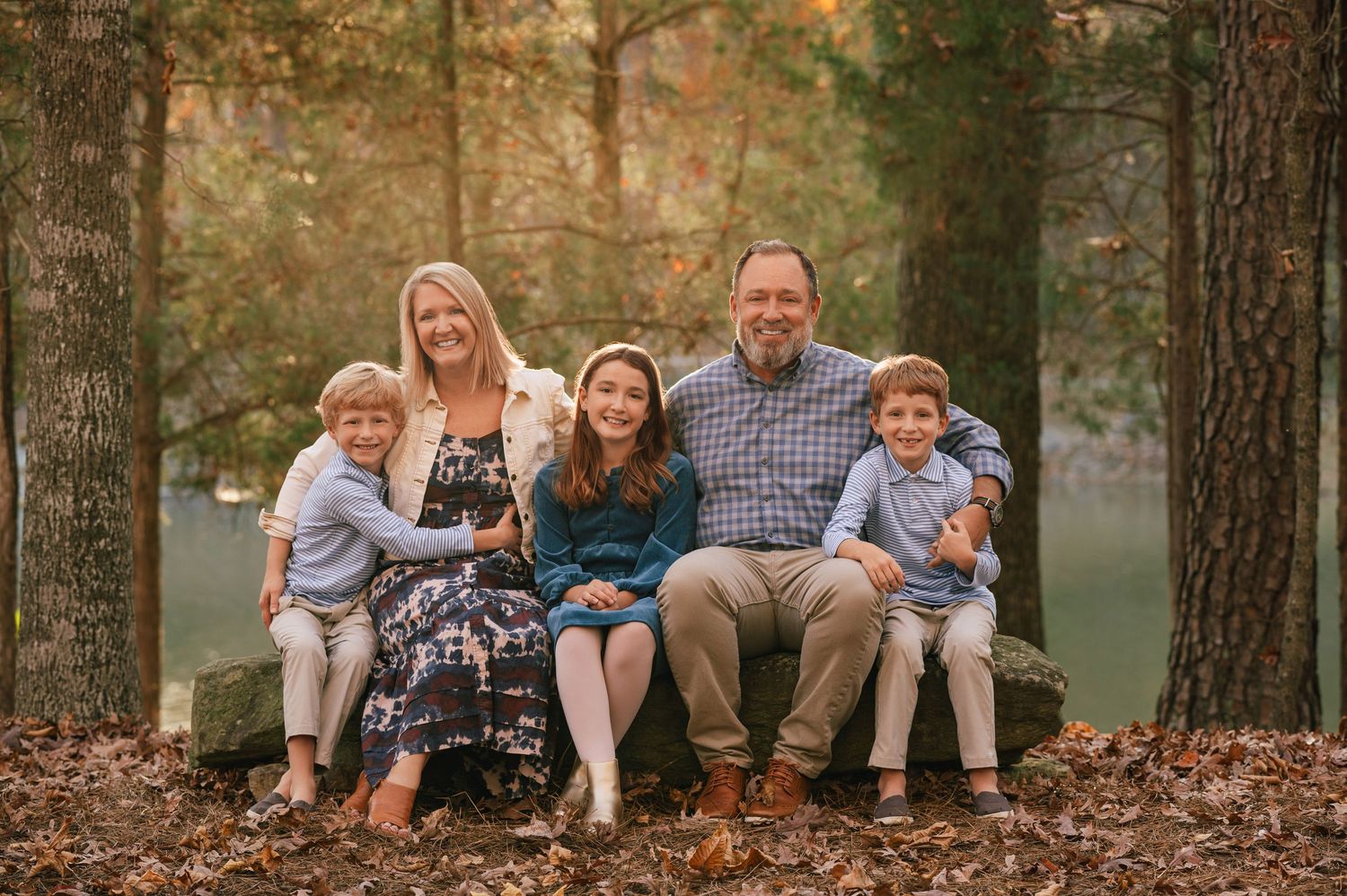 Family of five smiling outdoors in autumn forest setting with golden foliage, seated on rocks near a pond.