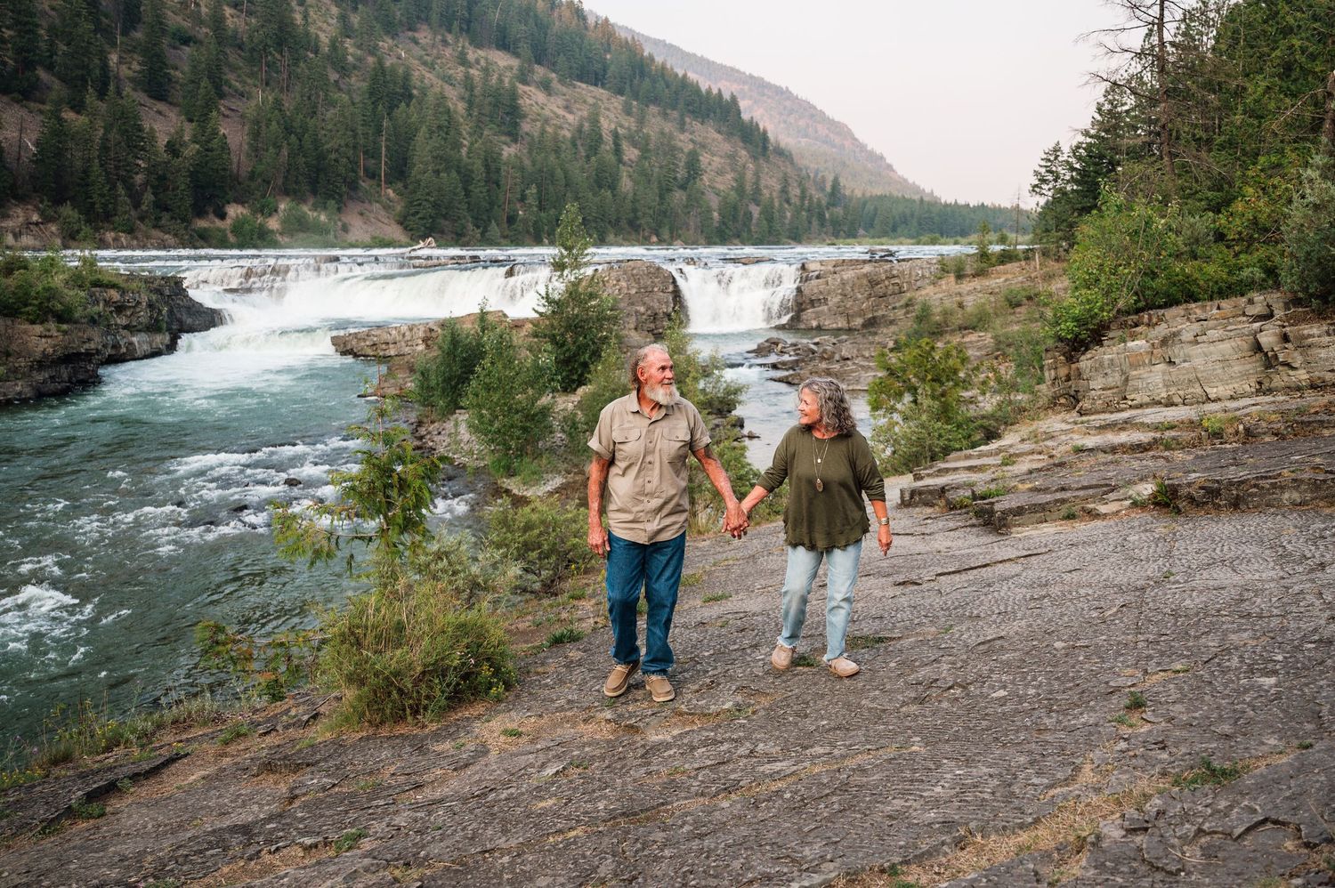 Couple holding hands on rocky riverbank with waterfall and evergreen forest backdrop in Pacific Northwest.