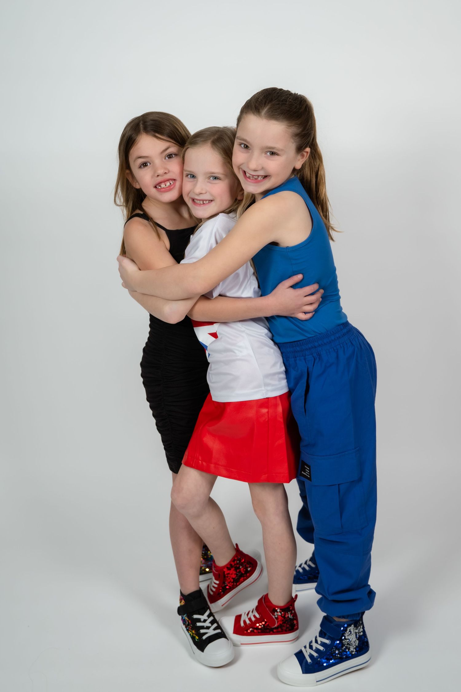 Three young girls hugging and smiling together in a studio, wearing colorful outfits and sneakers.