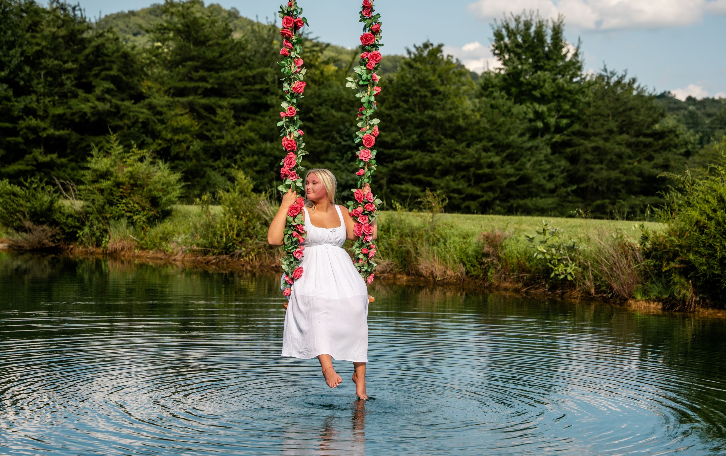 Woman in white dress holding floral swing over serene lake with lush green trees in background.