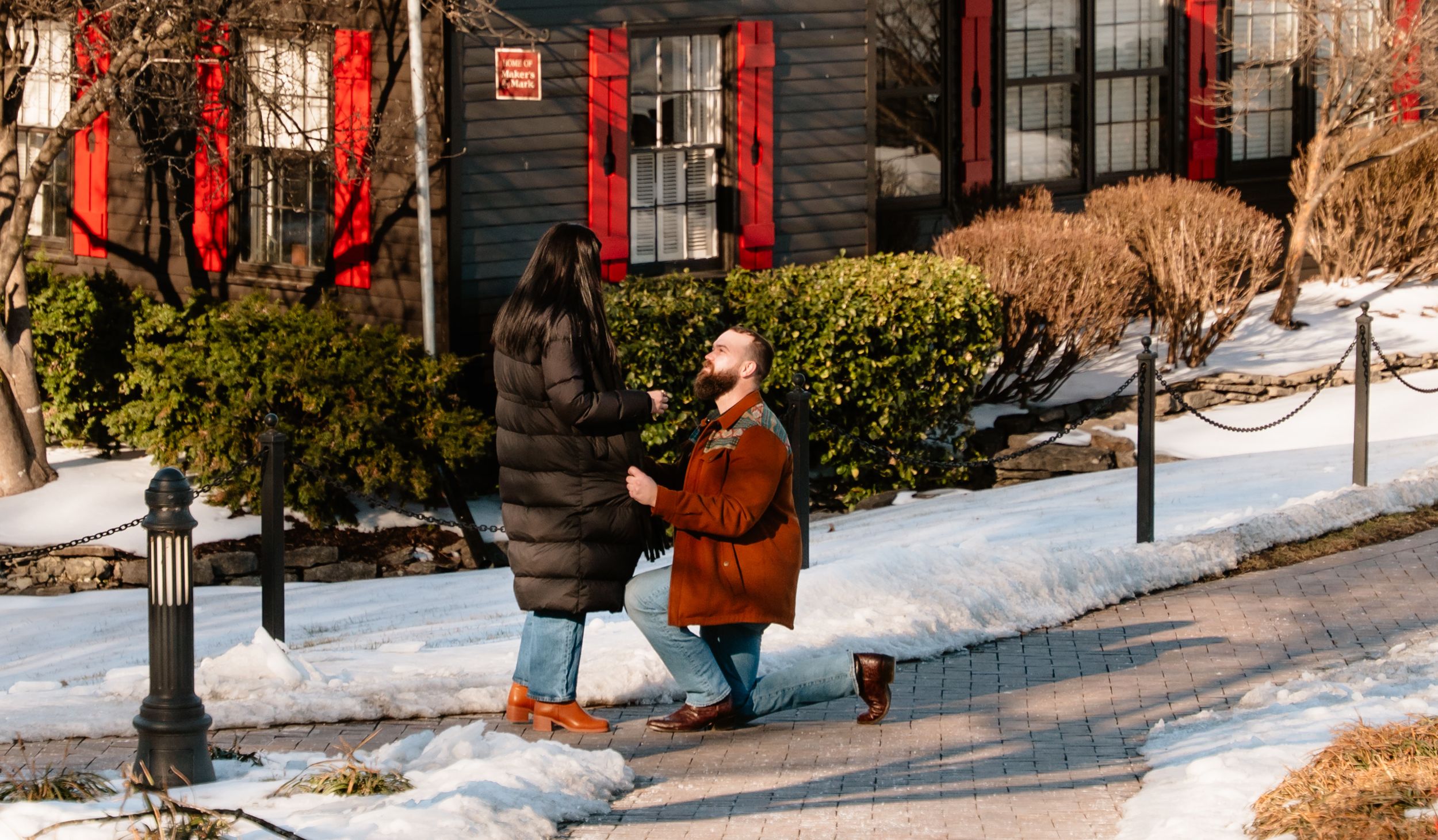 Man proposing on one knee to woman on snowy sidewalk in winter, red building backdrop, romantic outdoor proposal.
