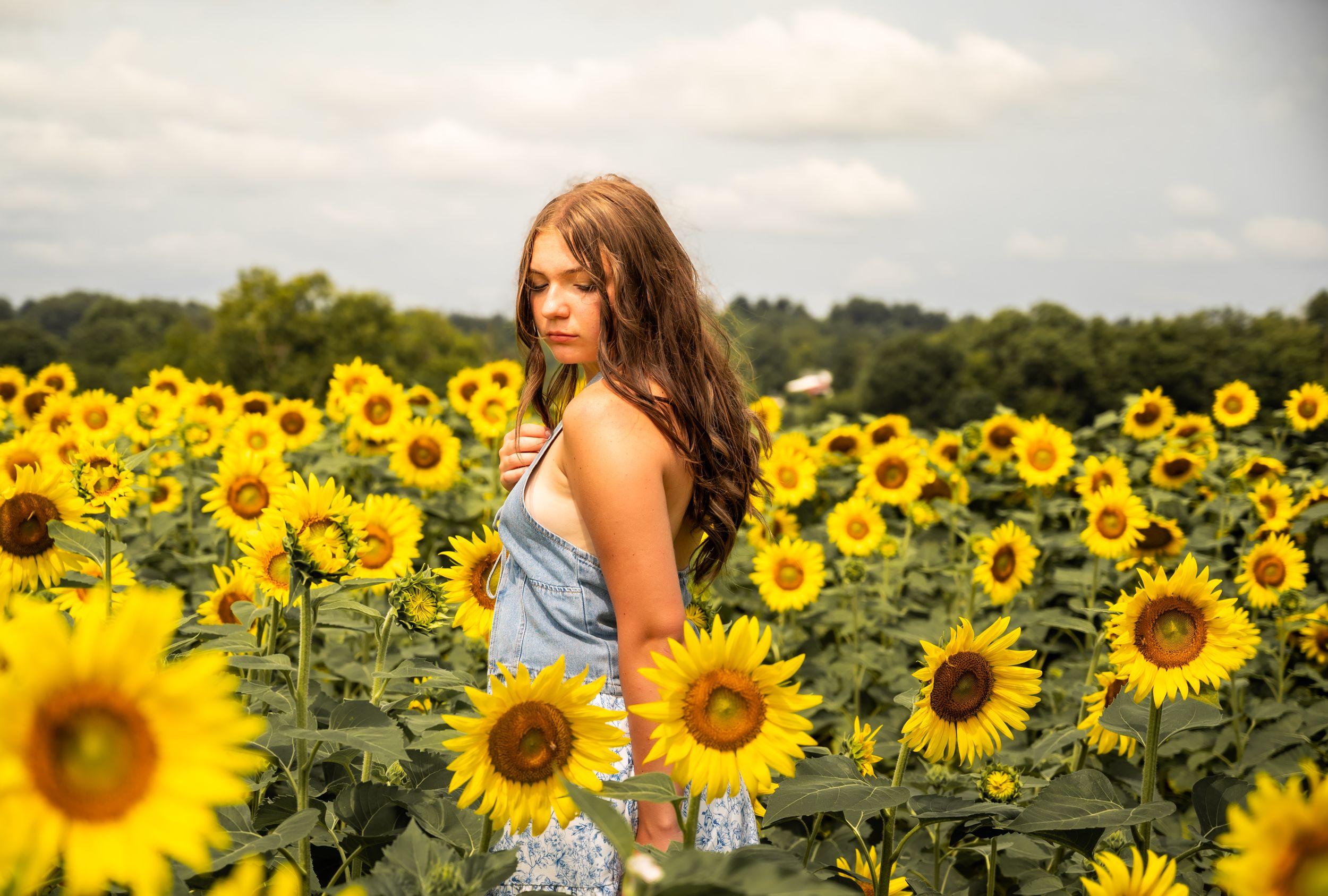 Young woman with long brown hair standing in a vibrant sunflower field on a cloudy day.