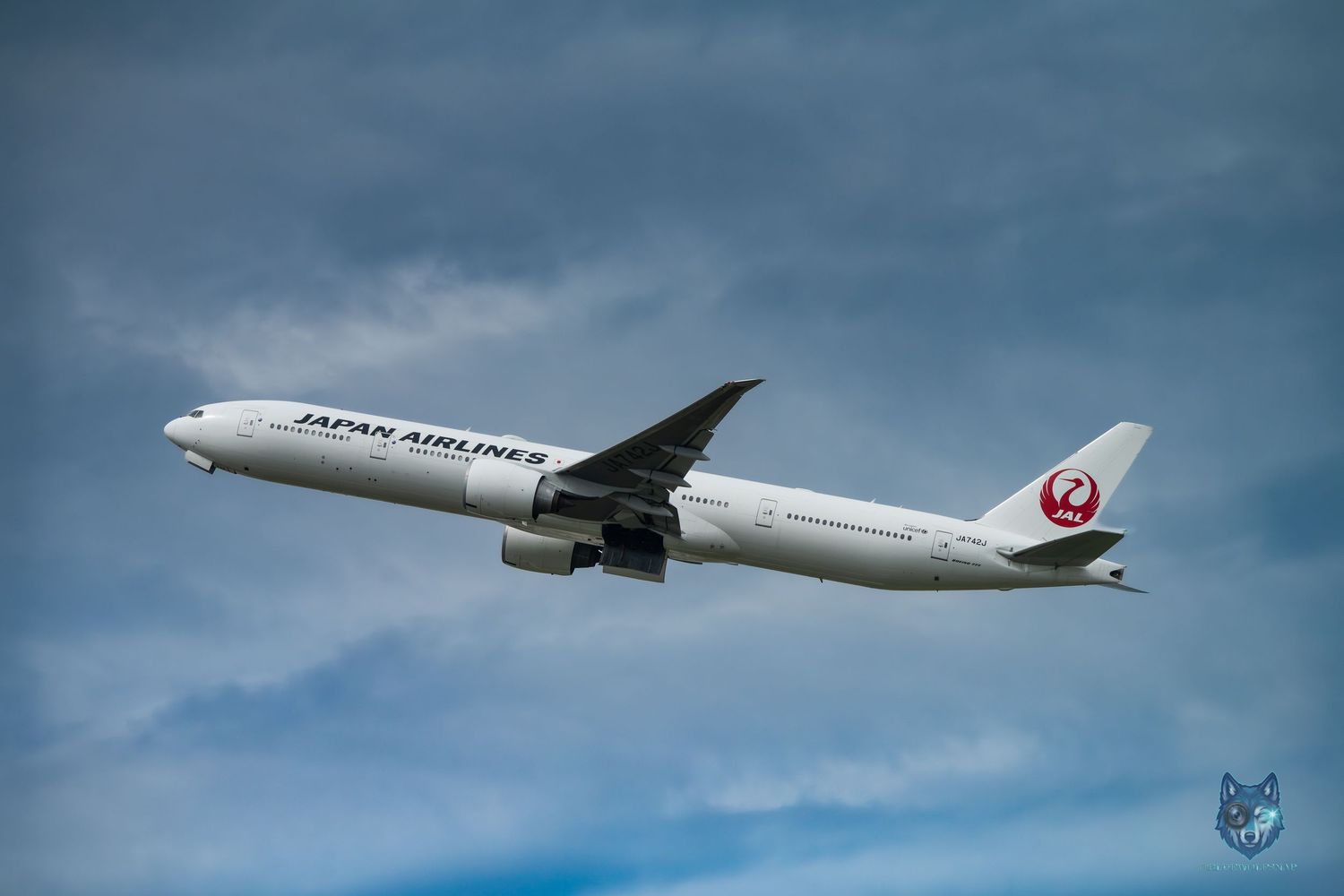 Japan Airlines Boeing 777 aircraft flying through cloudy blue skies, showcasing the iconic red crane logo on tail.
