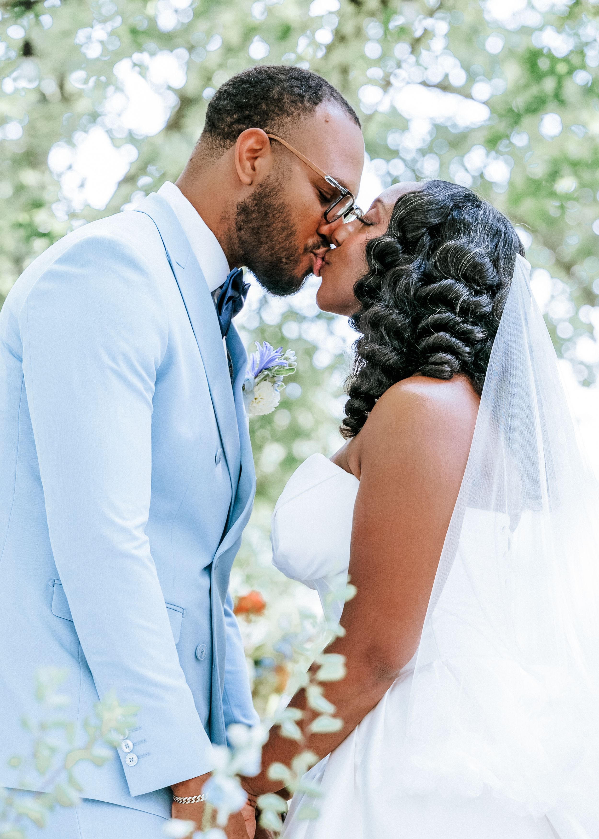 A Black bride and groom share a kiss outdoors, she in a white gown with veil, he in a light blue suit.