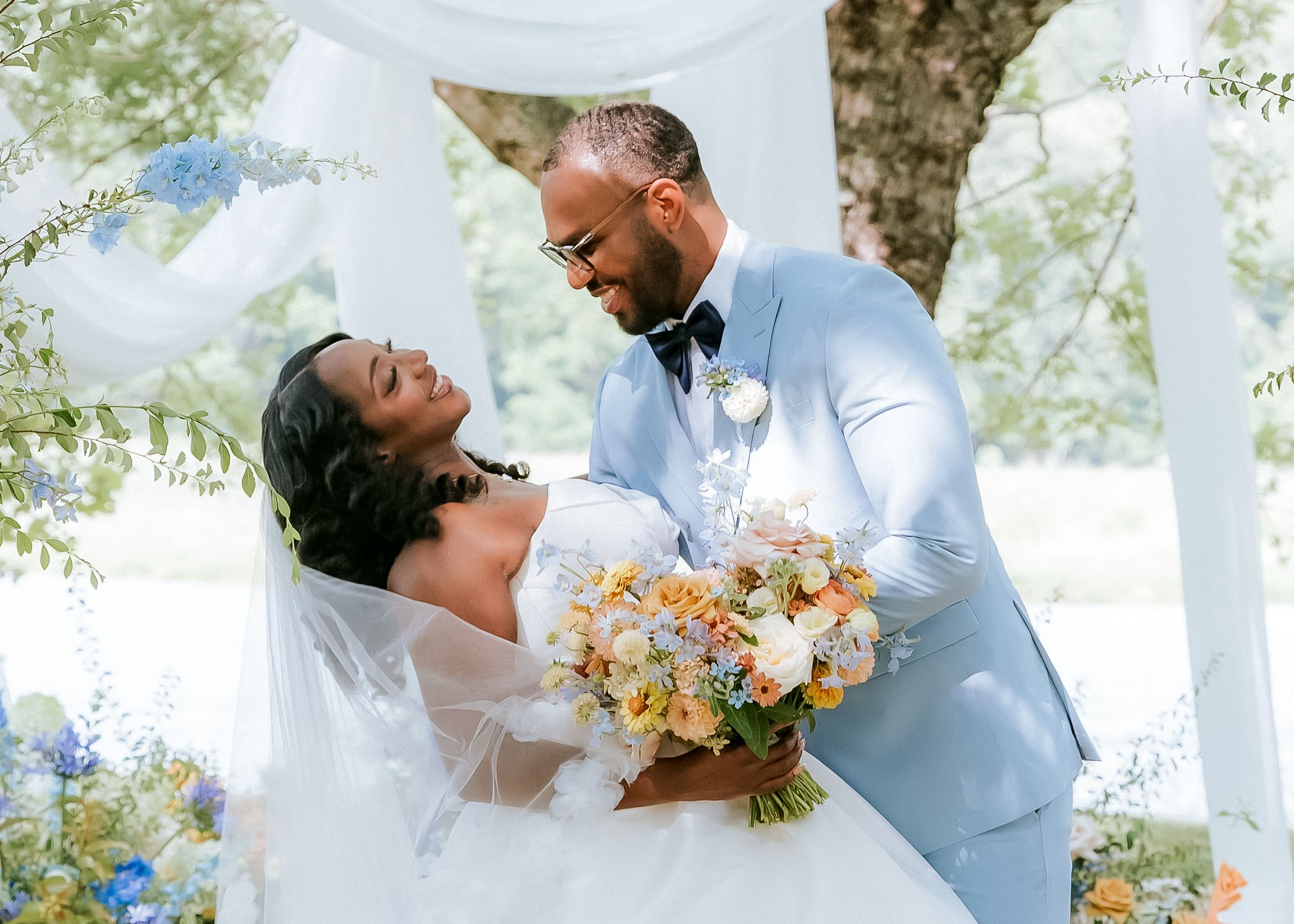 A bride and groom share a romantic moment under a floral arch, with the bride holding a colorful bouquet outdoors.