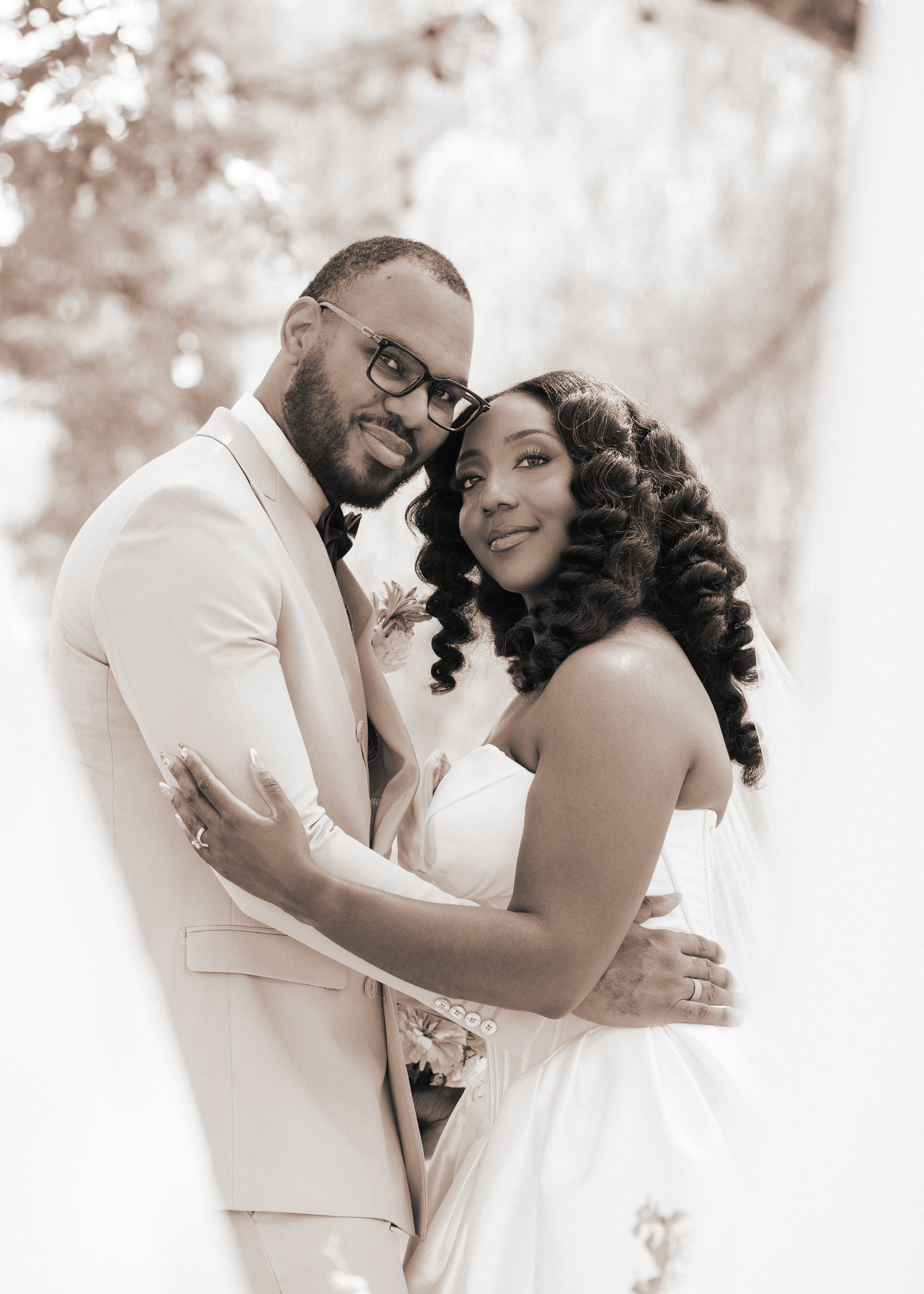 A Black couple poses in wedding attire, groom in white suit and bride in strapless gown, in a romantic sepia-toned portrait.