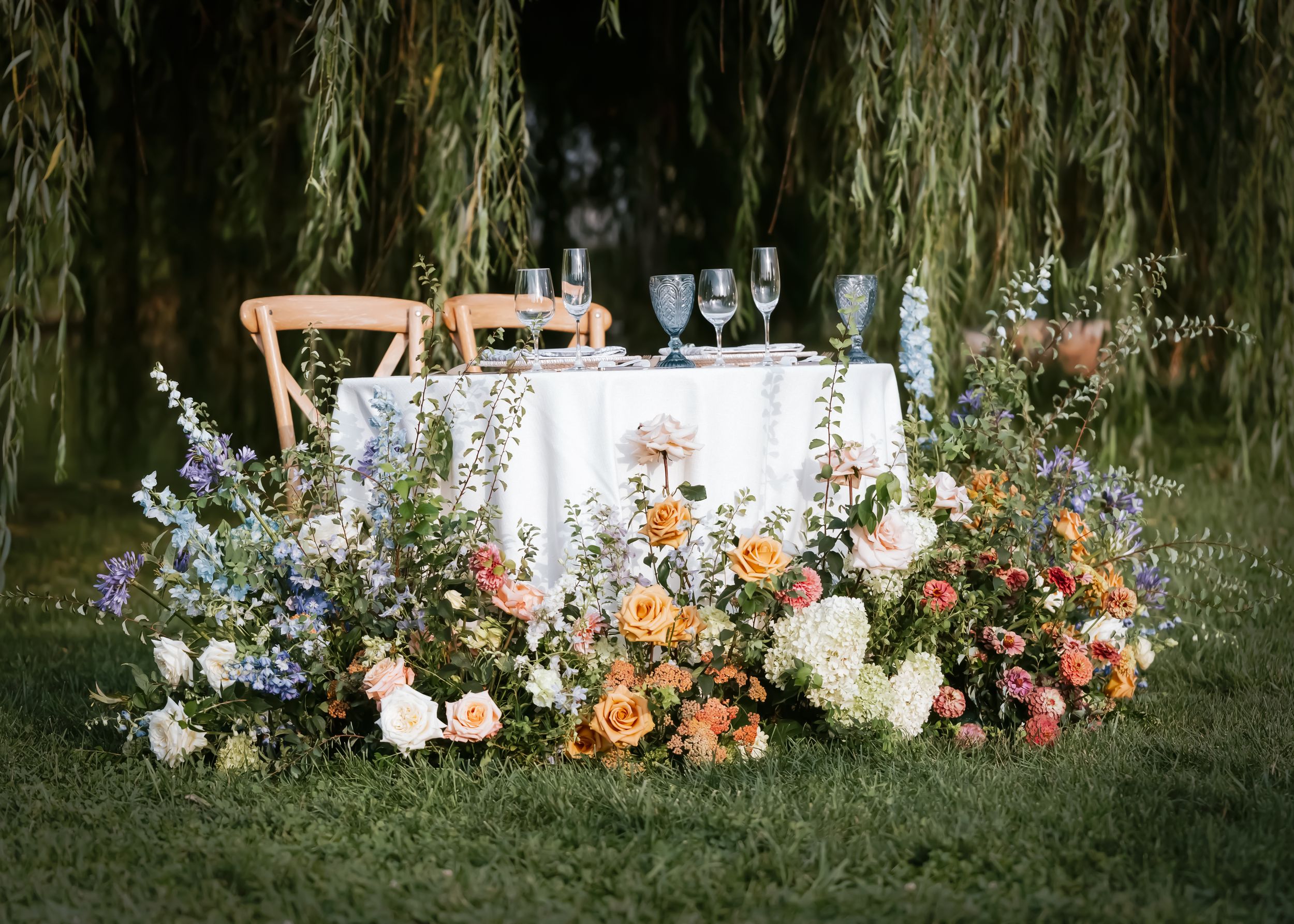 Outdoor sweetheart table with lush floral arrangements in peach, pink, and blue tones beneath weeping willow trees.