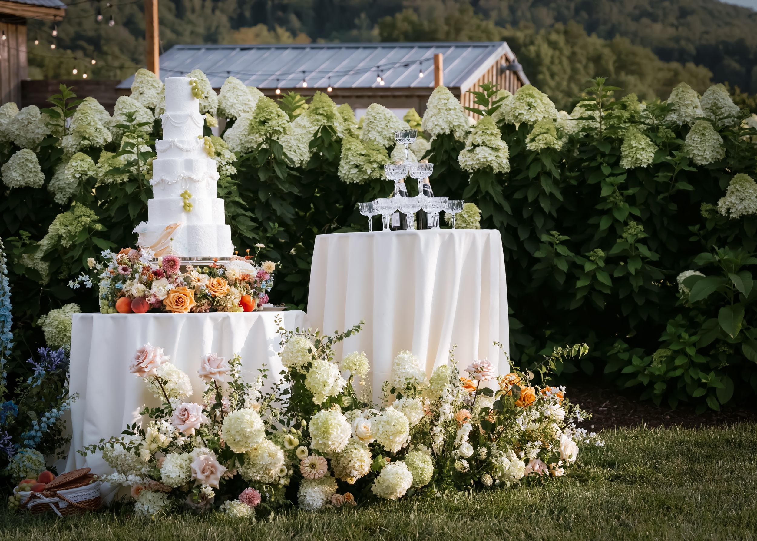 Elegant outdoor wedding cake display with white tiered cake, champagne tower, and lush floral arrangements on green lawn.