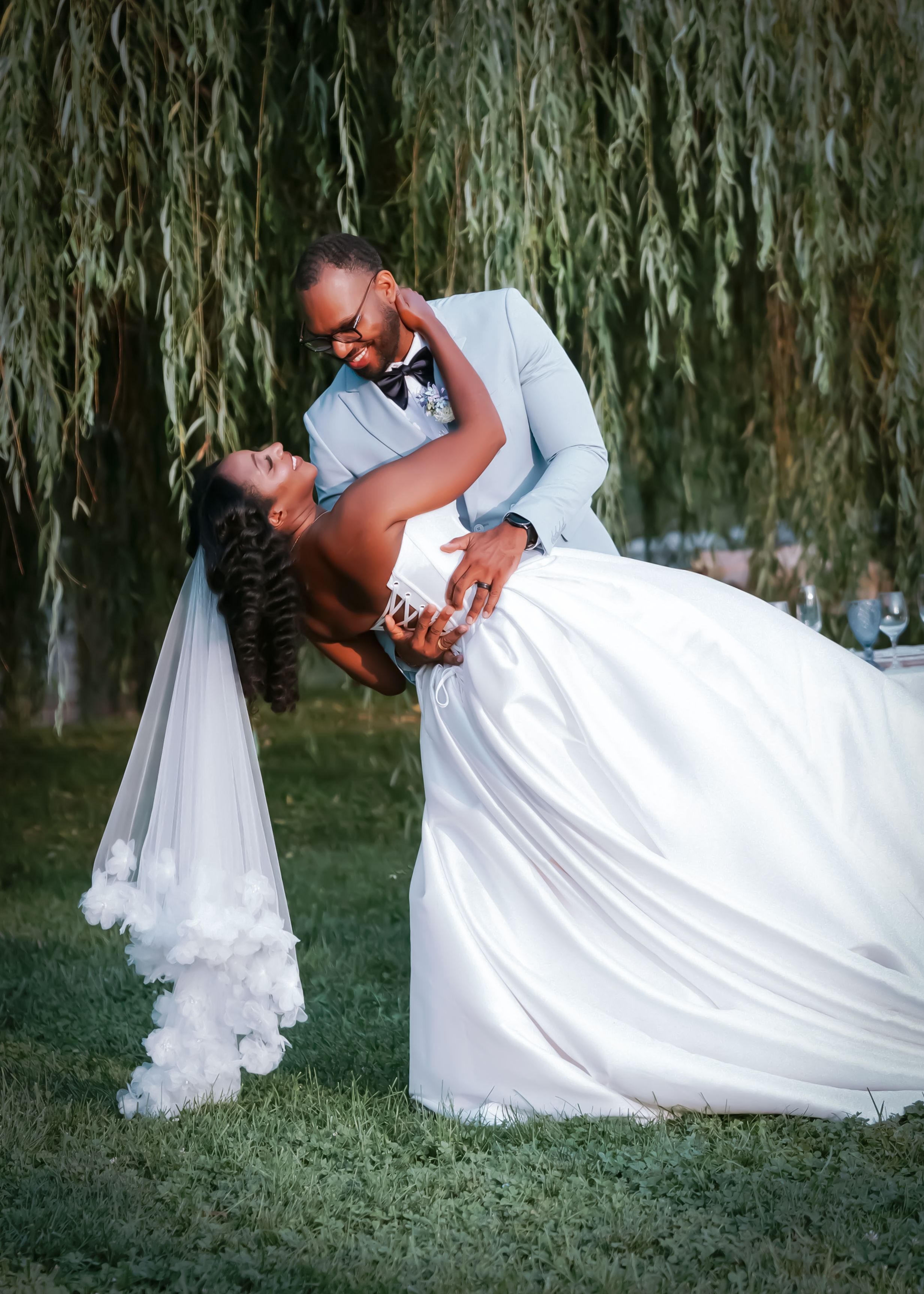 Bride in white gown and groom in light blue suit share a romantic dip kiss under a weeping willow tree.