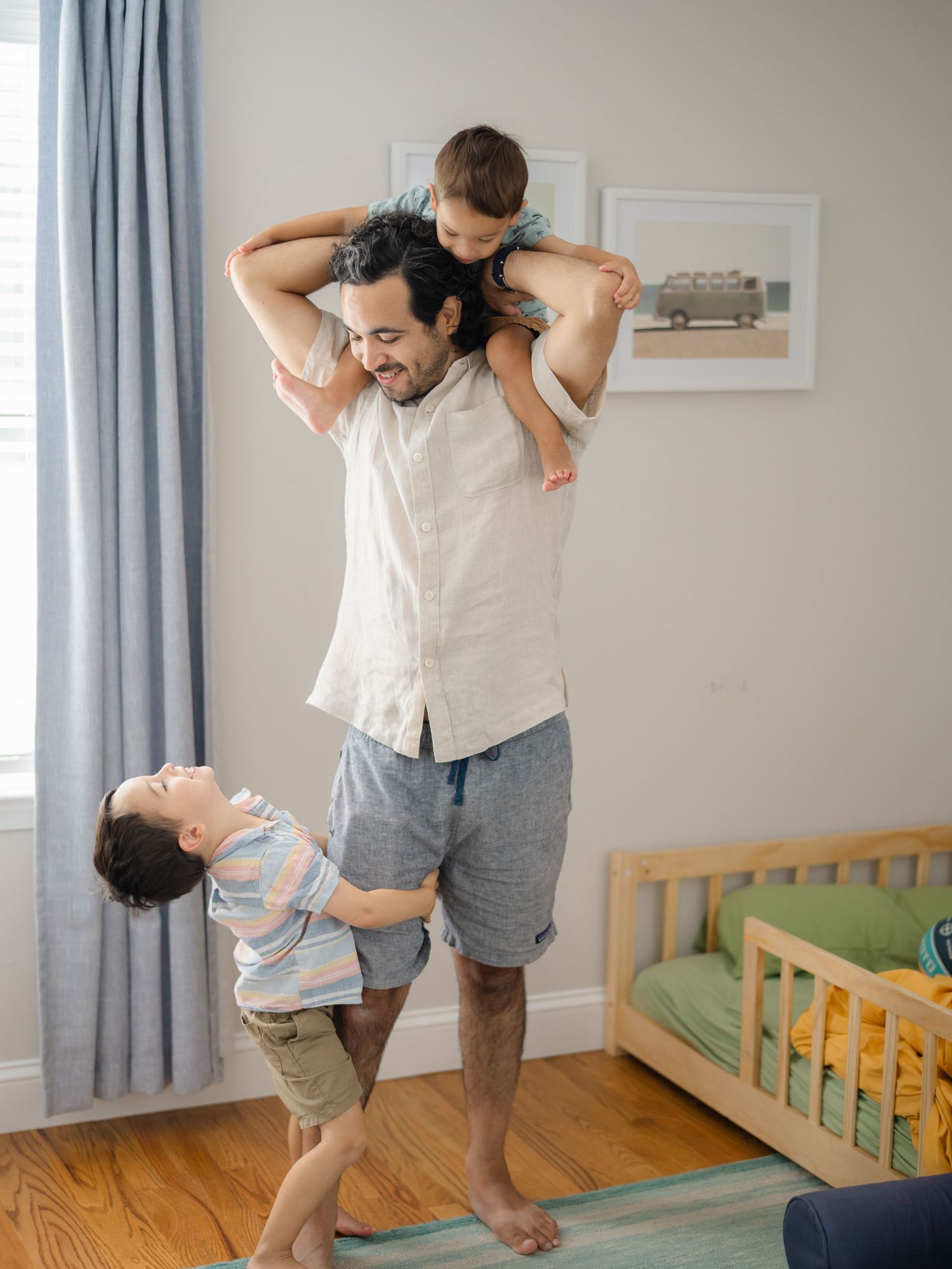 Kids climb on dad in brother's room with a window behind them.